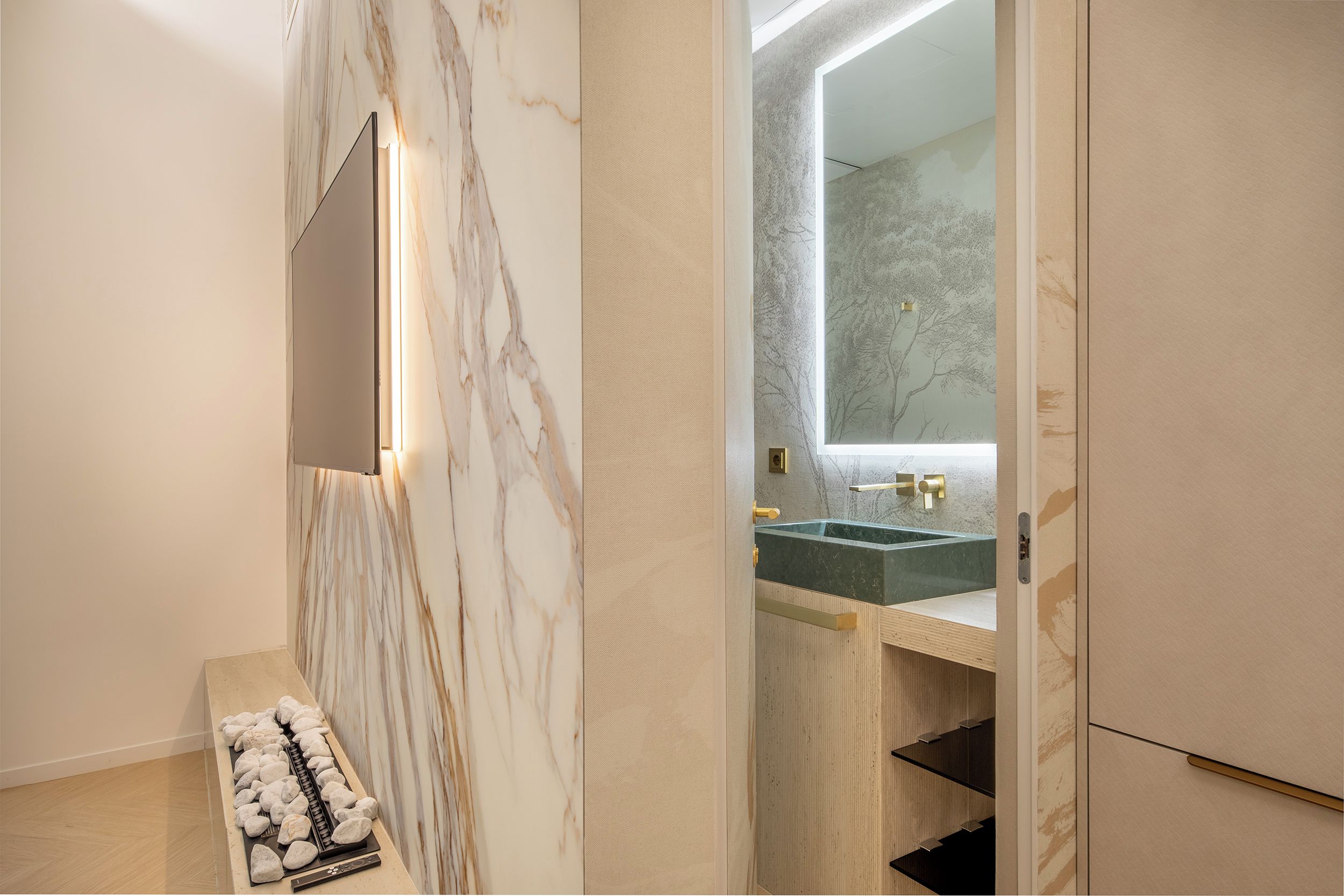 A modern bathroom with a marble wall, a large mirror, a sink with a gold faucet, and a bench with a towel.