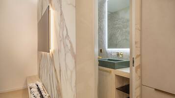 A modern bathroom with a marble wall, a large mirror, a sink with a gold faucet, and a bench with a towel.