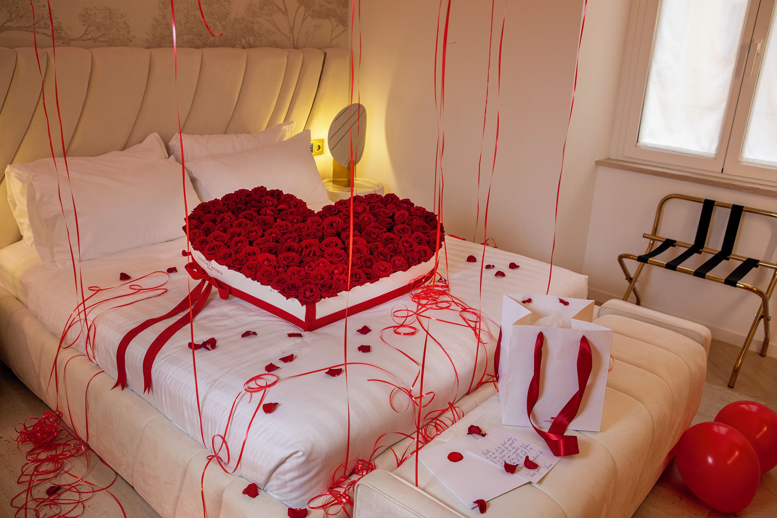 A bed with a heart-shaped bouquet of red roses, white pillows, and scattered rose petals.