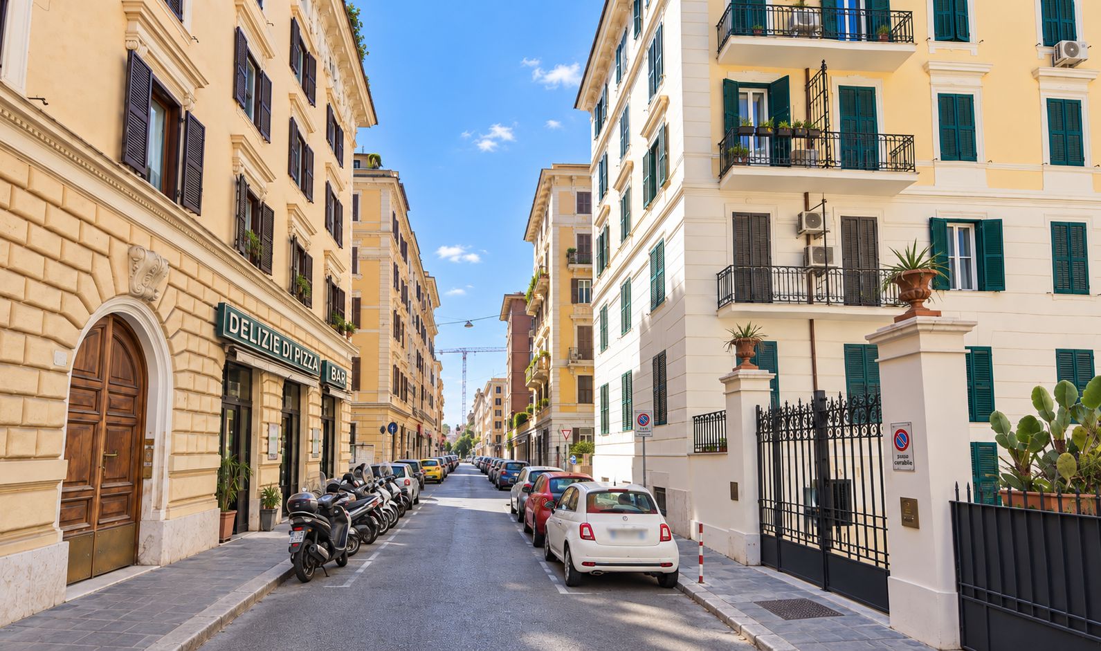 Una calle con coches aparcados y un hilera de edificios con balcones, un cartel de un bar de pizza, y un cartel de prohibido aparcar.