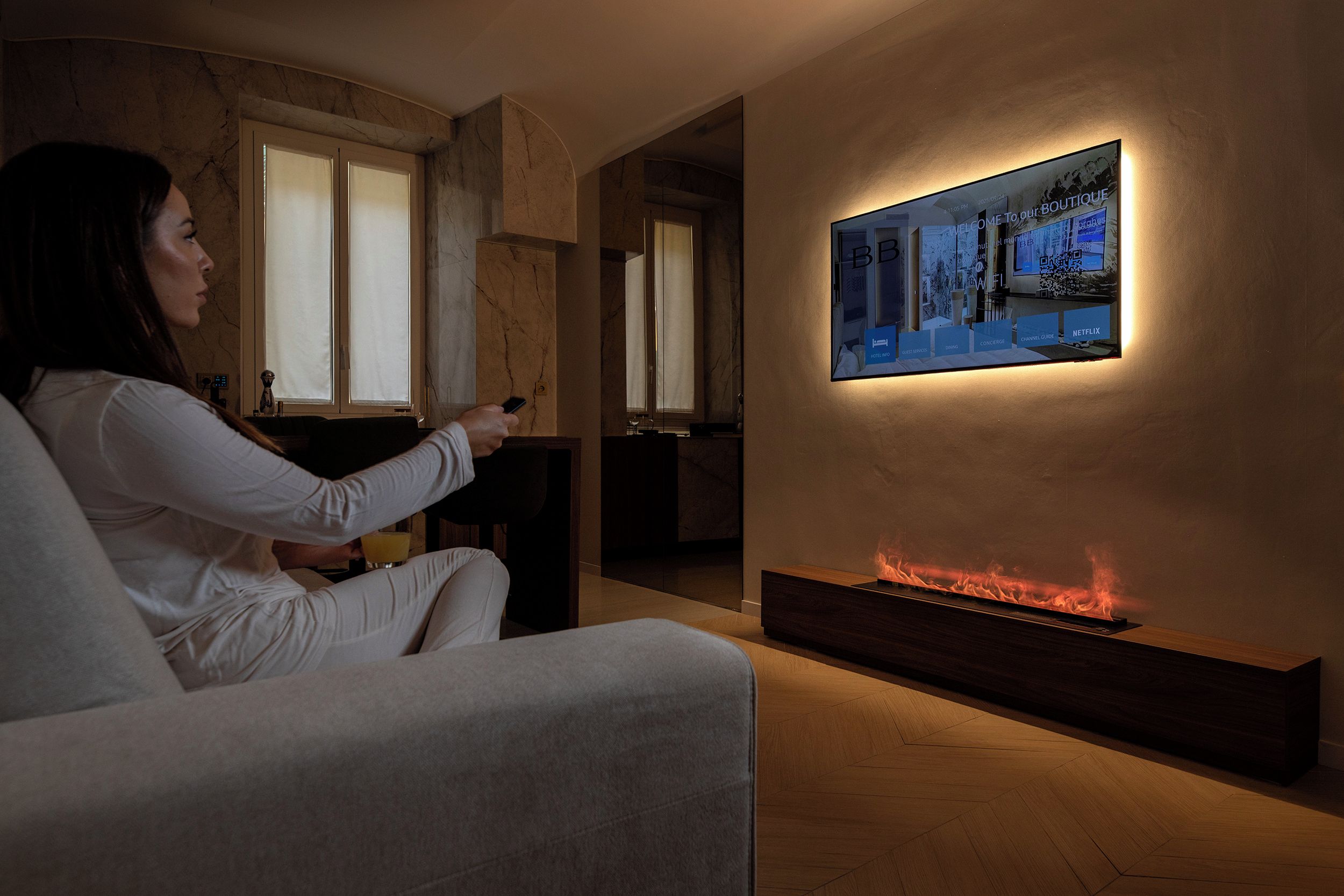 Una persona sentada en un sofá en una habitación con chimenea, un televisor de pared mostrando el interior de un hotel y una ventana con persianas.