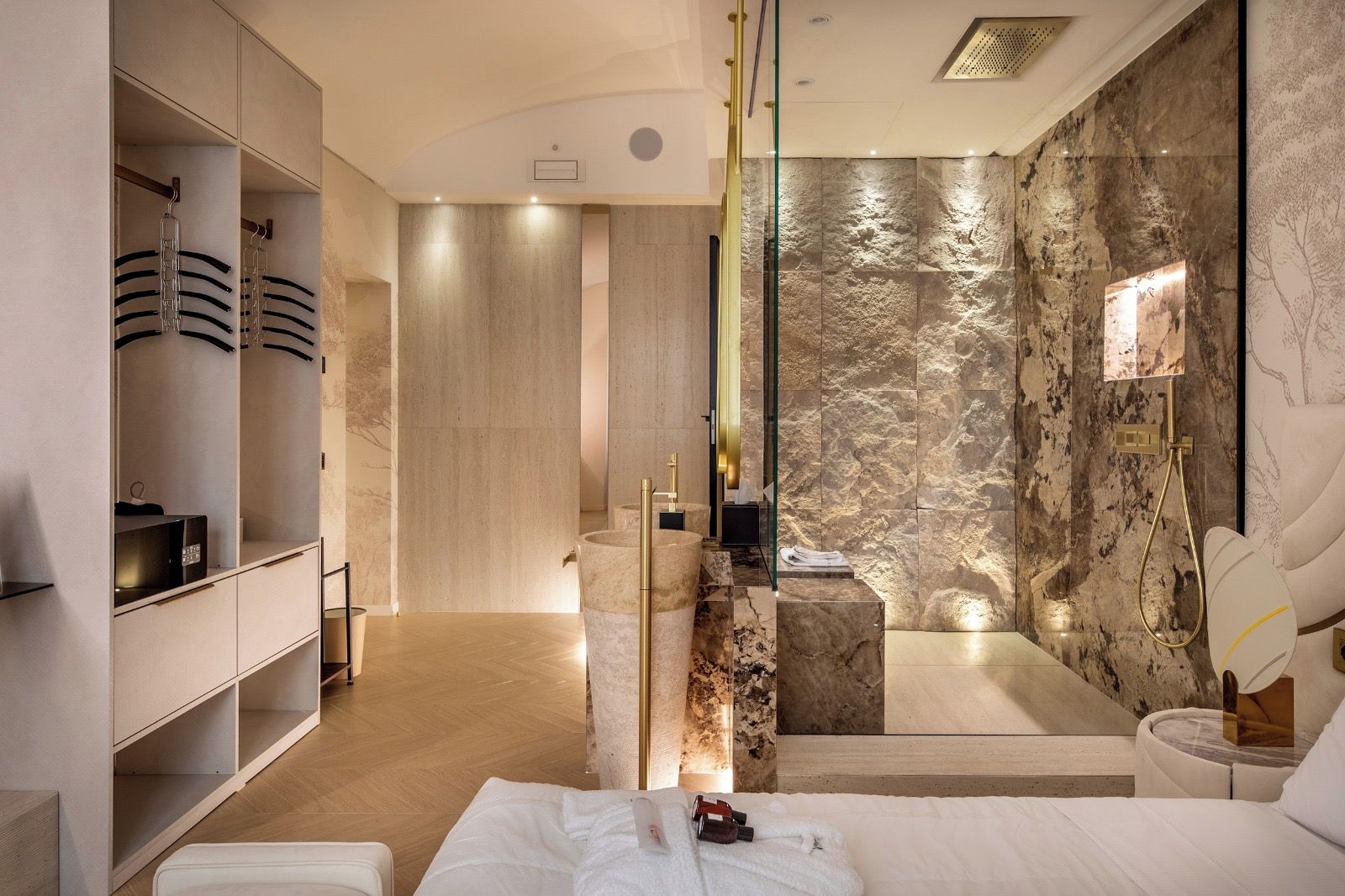 Una habitación de hotel moderna con una cama grande, un vestidor, un baño con pared de mármol y zona de ducha.