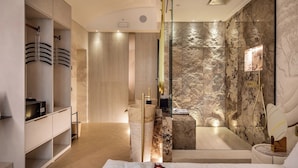 Una habitación de hotel moderna con una cama grande, un vestidor, un baño con pared de mármol y zona de ducha.