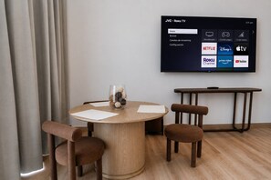 A modern living room with a round wooden table, two brown chairs, a glass vase with decorative items, and a flat-screen TV mounted on the wall displaying various streaming services. - Roma Residence | Luxury Suite 201 (Mexico City)