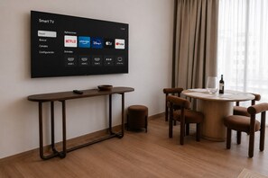 A modern hotel room with a large flat-screen TV displaying a smart TV interface, a round dining table with chairs, a small stool, and a wine glass on the table. - Roma Residence | Luxury Suite 203 (Mexico City)