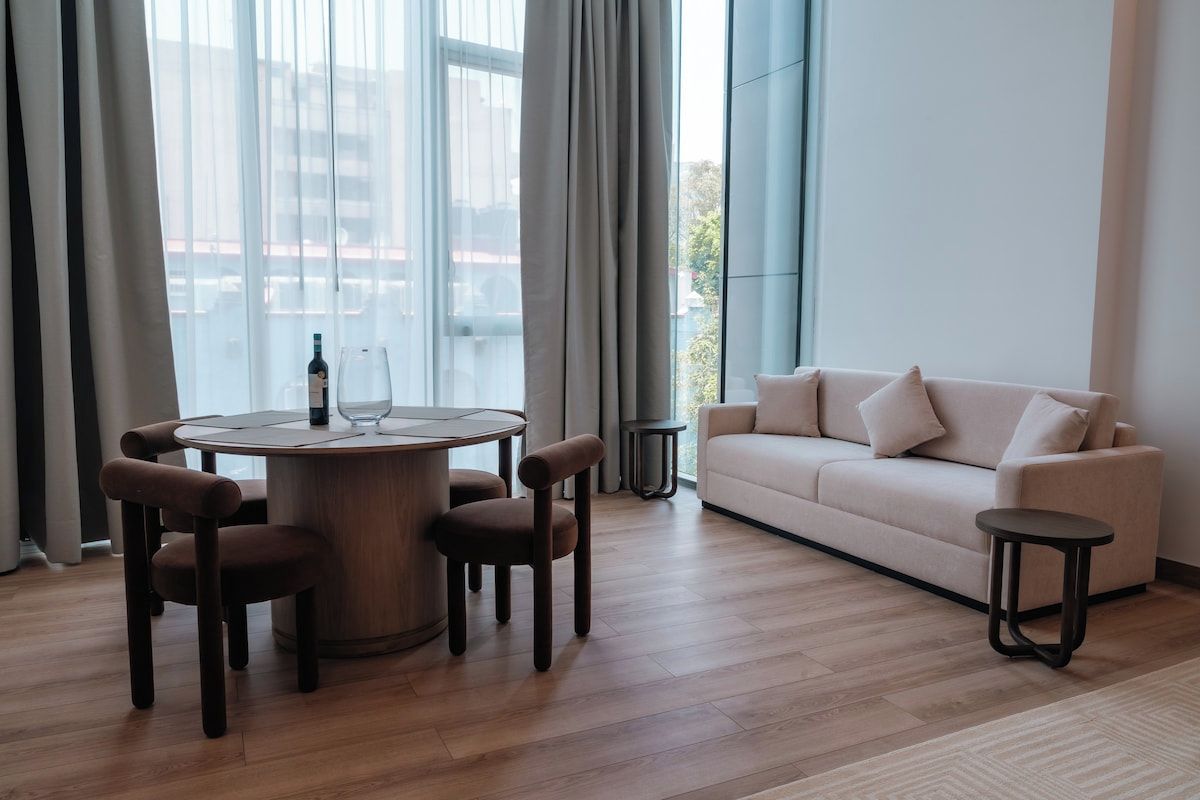 En moderne stue med et rundt træbord, fire brune stole, en beige sofa og to små sorte sofaborde.