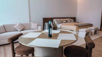 A modern hotel room with a round wooden table, two brown chairs, a white sofa, and a bed with a wooden headboard.