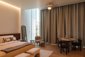 Une chambre d’hôtel comprenant un lit, un bureau, deux chaises, une table ronde et une grande fenêtre avec des rideaux.