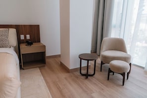 Ein Hotelzimmer mit einem Bett, einem Nachttisch mit Telefon, einem runden Tisch, einem Sessel und Blick ins Freie durch große Fenster.