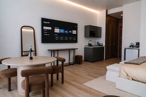 A modern hotel room with a round dining table, brown upholstered chairs, a flat-screen TV mounted on the wall, a kitchenette, and a bed with a white headboard. - Condesa Residence - Roma Luxury Suite 302 (Mexico City)