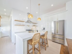 A modern kitchen with white cabinetry, a central island with bar stools, a stainless steel refrigerator, and pendant lighting. - Modern Condo Above Boutiques + Beach Nearby (Cape May)