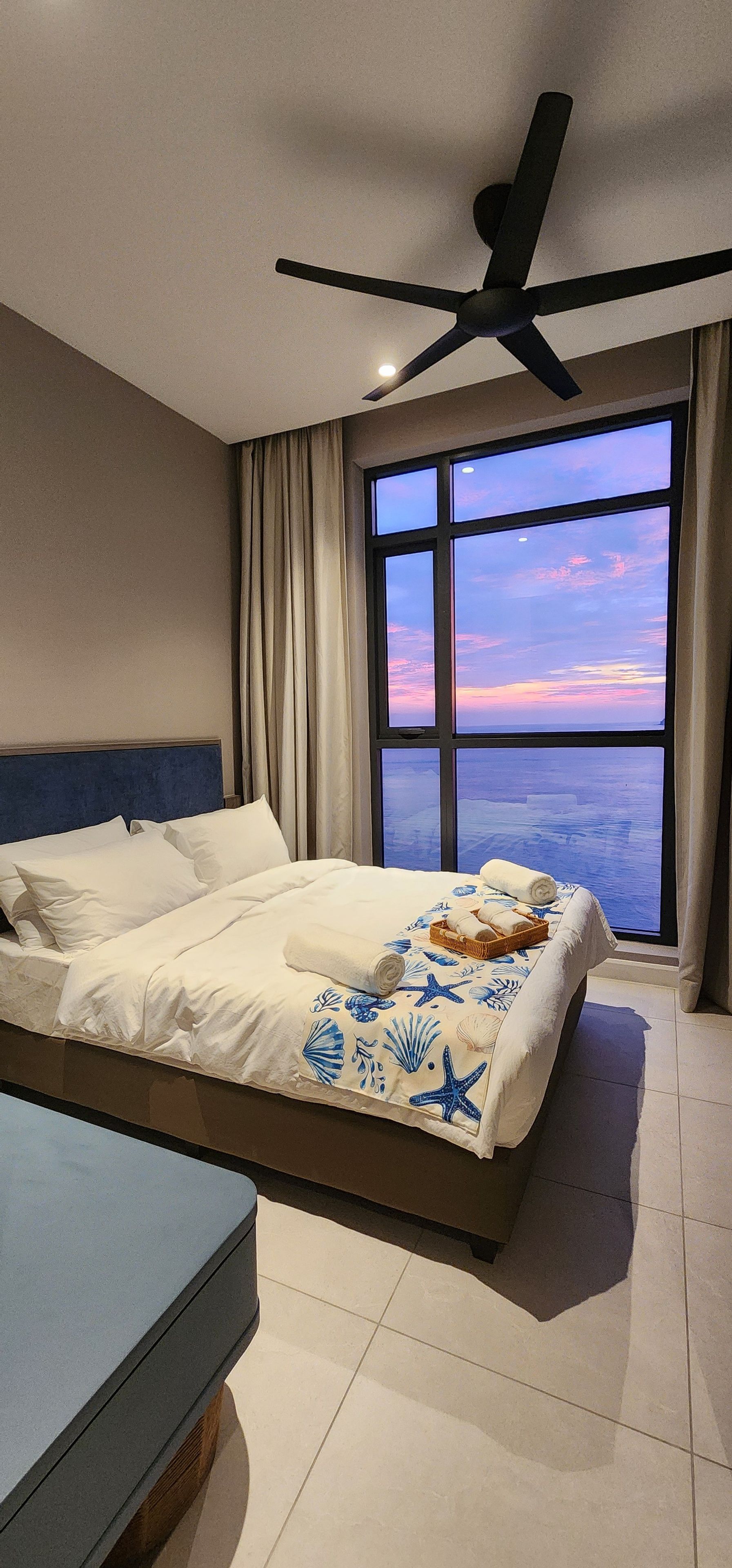 Ein modernes Hotelzimmer mit einem großen Bett, einem Deckenventilator und Blick auf den Ozean bei Sonnenuntergang.
