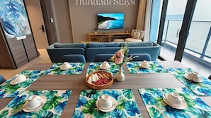 Un coin repas avec une grande table dressée pour le service du thé, un téléviseur en arrière-plan et une vue sur la mer par les fenêtres.