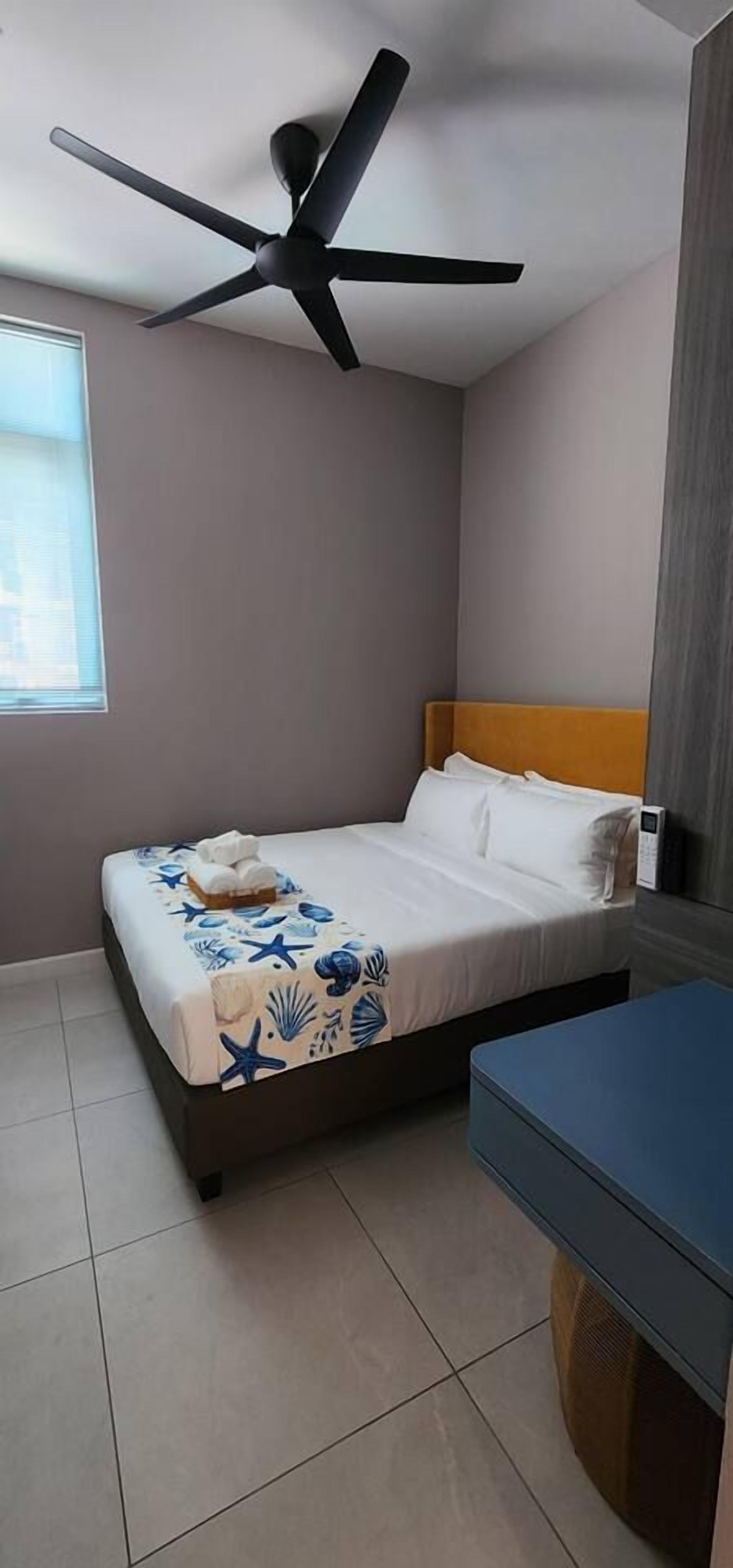 A hotel room with a bed, a ceiling fan, a bedside table, and a window.