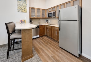 A compact kitchen with wooden cabinets, a stainless steel refrigerator, a microwave, and a dining area with a chair and table.