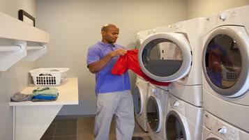 A laundry room with stacked washing machines, a laundry basket, and a shelf with folded clothes.