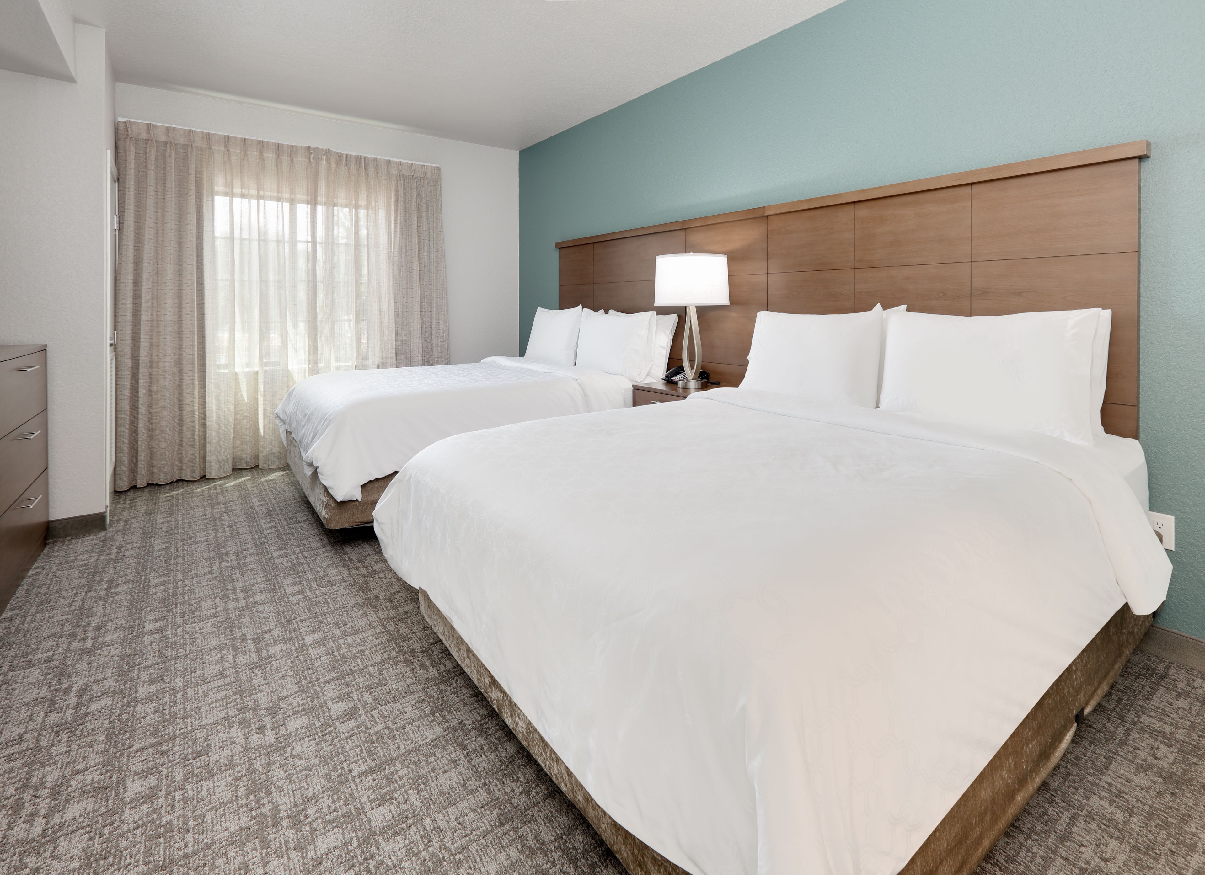 Ein Hotelzimmer mit zwei Betten, einem Nachttisch mit Lampe, einem Schminktisch und einem Fenster mit Vorhängen.