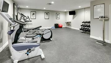 Sala de ginástica com esteiras, bicicleta ergométrica, uma bola de exercício vermelha e um suporte com halteres.