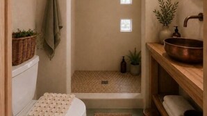 Casa de banho com sanita branca, toucador de madeira com lavatório de cobre, uma pequena planta em vaso e uma toalha pendurada na parede.