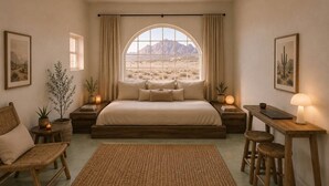 Quarto com cama grande, escrivaninha, duas cadeiras, janela com vista para o deserto e pequena mesa com abajur.