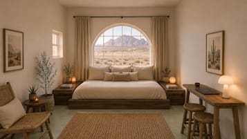 A bedroom with a large bed, a desk, two chairs, a window with a desert view, and a small table with a lamp.