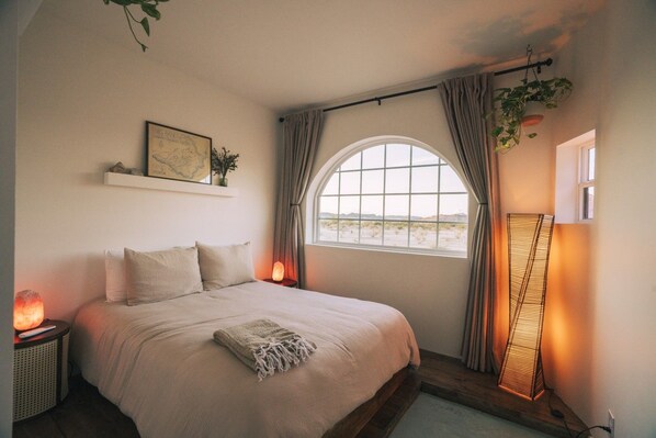 A bedroom with a large bed, a window with curtains, a wall-mounted shelf with a map, and a floor lamp.