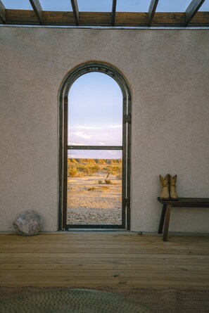 A room with a wooden floor, a bench, a pair of boots, and a large arched doorway leading to a landscape view.