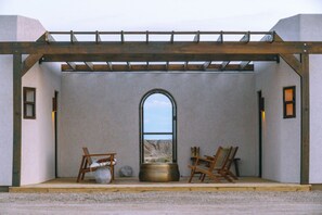 A room with a view through an arched window, a wooden pergola, and two chairs with a pot and a vase on the floor.