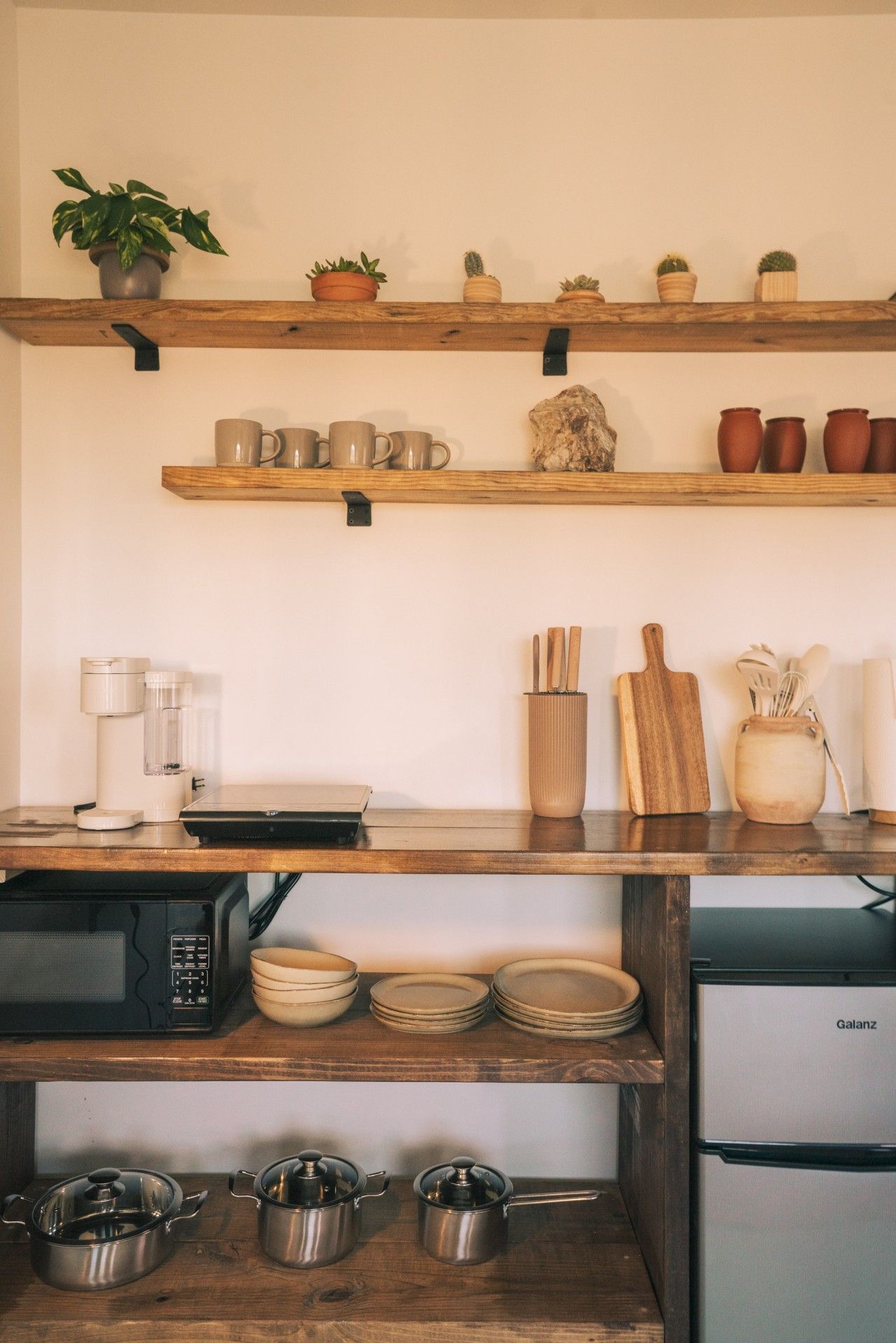 A kitchen with wooden shelves holding mugs, a coffee maker, a cutting board, and a Galanz refrigerator.