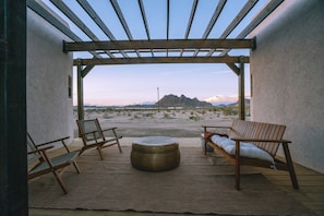 En udendørs terrasse med et pergolaoverdækket område, to træstole, et rundt bord og udsigt til et bjerglandskab.