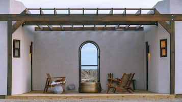 A room with a view through an arched window, a wooden pergola, and two chairs with a pot and a vase on the floor.