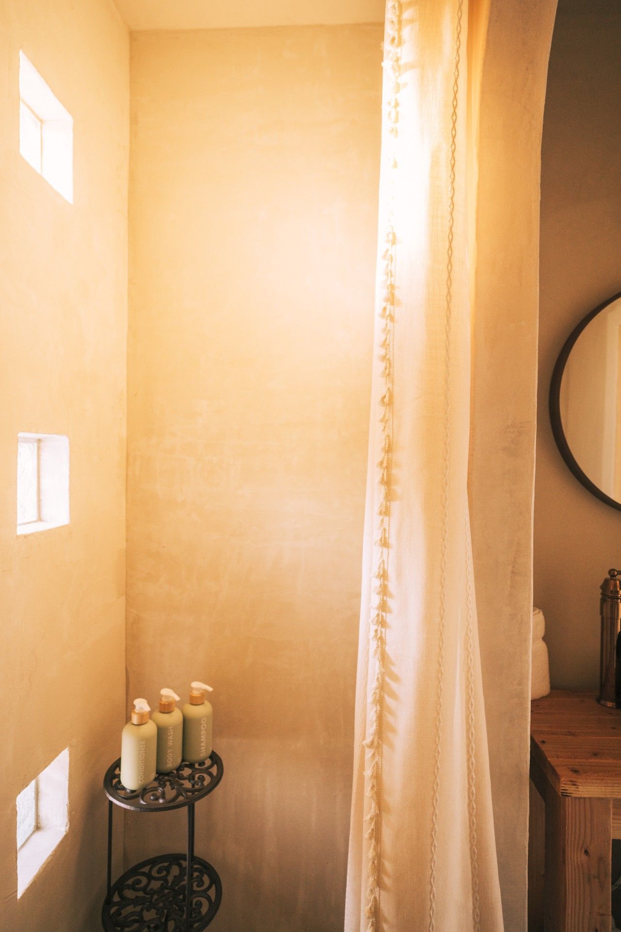 Un baño con pared beige, un mueble de madera, un espejo redondo y una mesita con tres botellas.