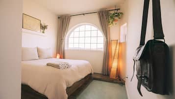 A bedroom with a bed, a window with curtains, a hanging plant, and a backpack on the wall.