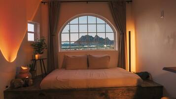 A bedroom with a large bed, a window with a view of mountains, a bedside table with a lamp, and a chair.