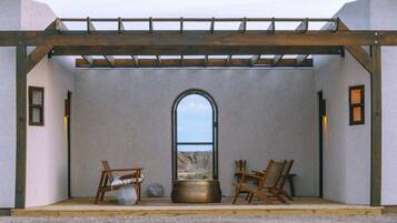 A room with a view through an arched window, a wooden pergola, and two chairs with a pot and a vase on the floor.