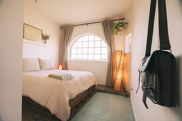 A bedroom with a bed, a window with curtains, a hanging plant, and a backpack on the wall.