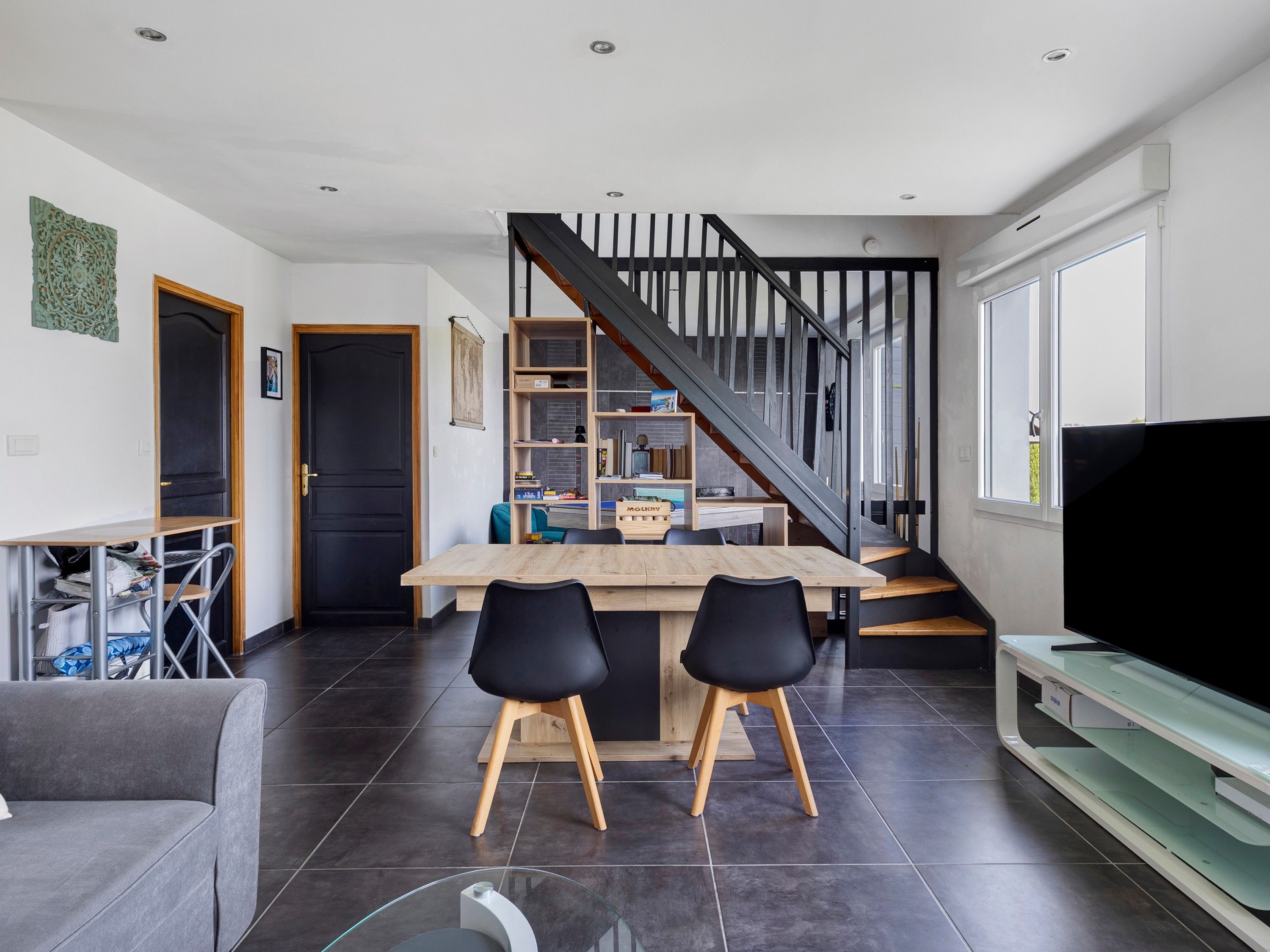 A modern living space with a kitchen area, a dining table with chairs, a staircase, and a flat-screen TV.