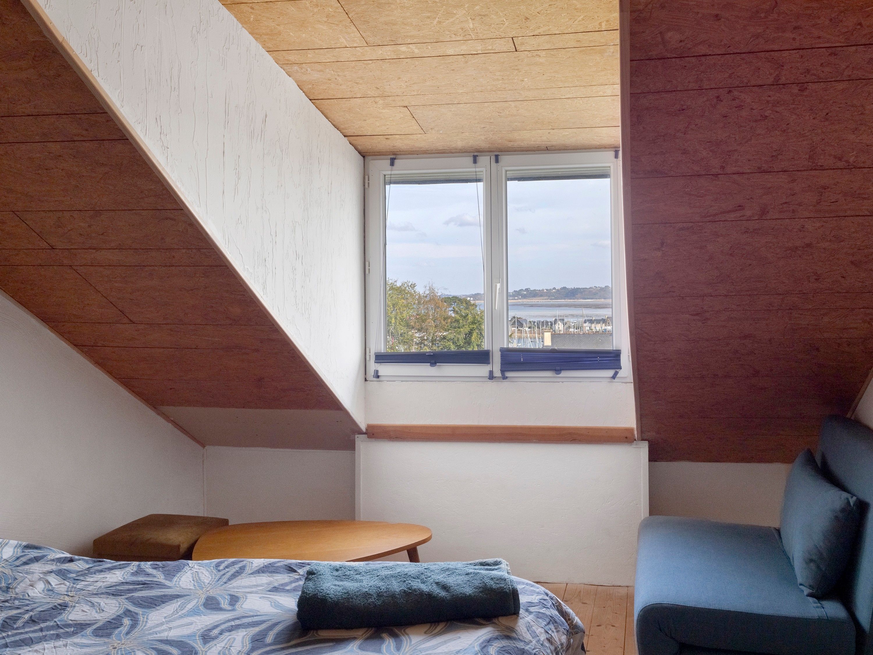 A bedroom with a sloped ceiling, a large window, a bed, a small table, and a blue chair.