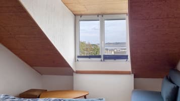 A bedroom with a sloped ceiling, a large window, a bed, a small table, and a blue chair.