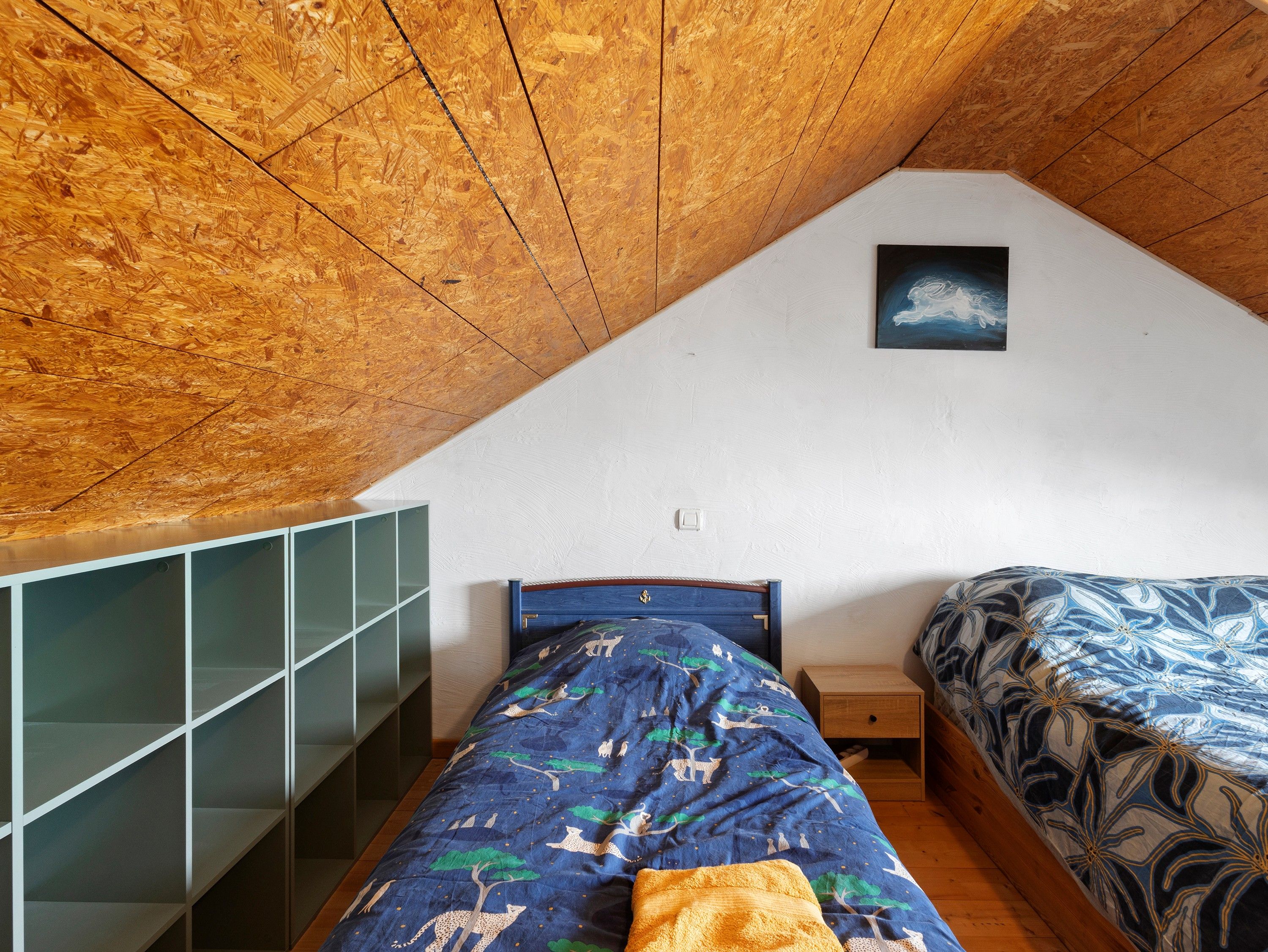 Un dormitorio con techo inclinado, una cama con colcha azul estampada, una mesita de noche y un estante montado en la pared.