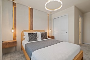 Ein ordentlich bezogenes Bett mit einem hölzernen Kopfteil, einer grauen Decke und gemusterten Kissen. Es befinden sich zwei Nachttische mit Lampen daneben, und im Hintergrund eine weiße Tür.