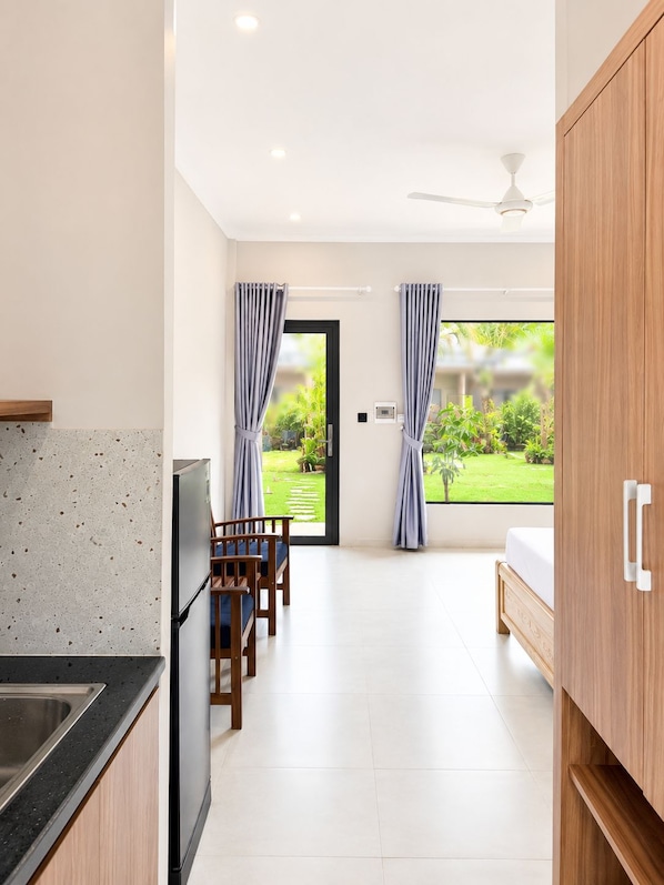 A modern kitchen with a countertop and cabinets, a dining area with chairs, a bed, and a view of a garden through a door.