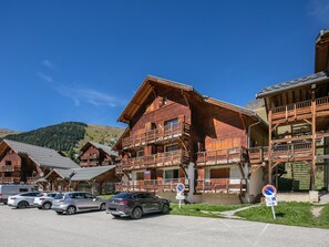 A row of wooden chalet-style buildings with balconies, a parking area with cars, and a clear blue sky. - Flat with a balcony and heated swimming pool / accessible for people with reduced mobility (Saint-Sorlin-d´Arves)