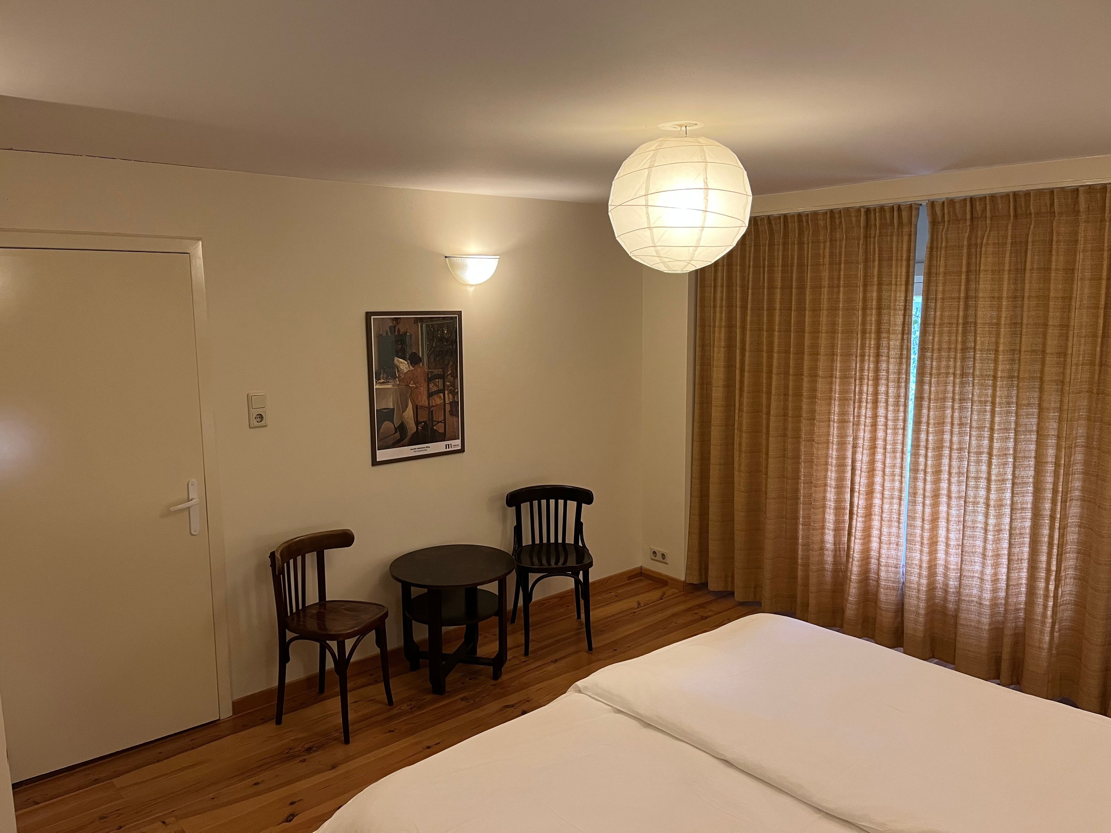Une chambre d’hôtel avec un lit, deux chaises, une petite table, une lampe fixée au mur, un tableau et un rideau.