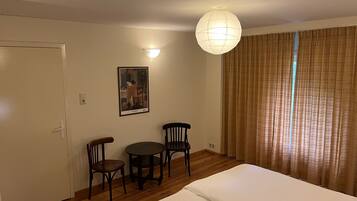 Une chambre d’hôtel avec un lit, deux chaises, une petite table, une lampe fixée au mur, un tableau et un rideau.