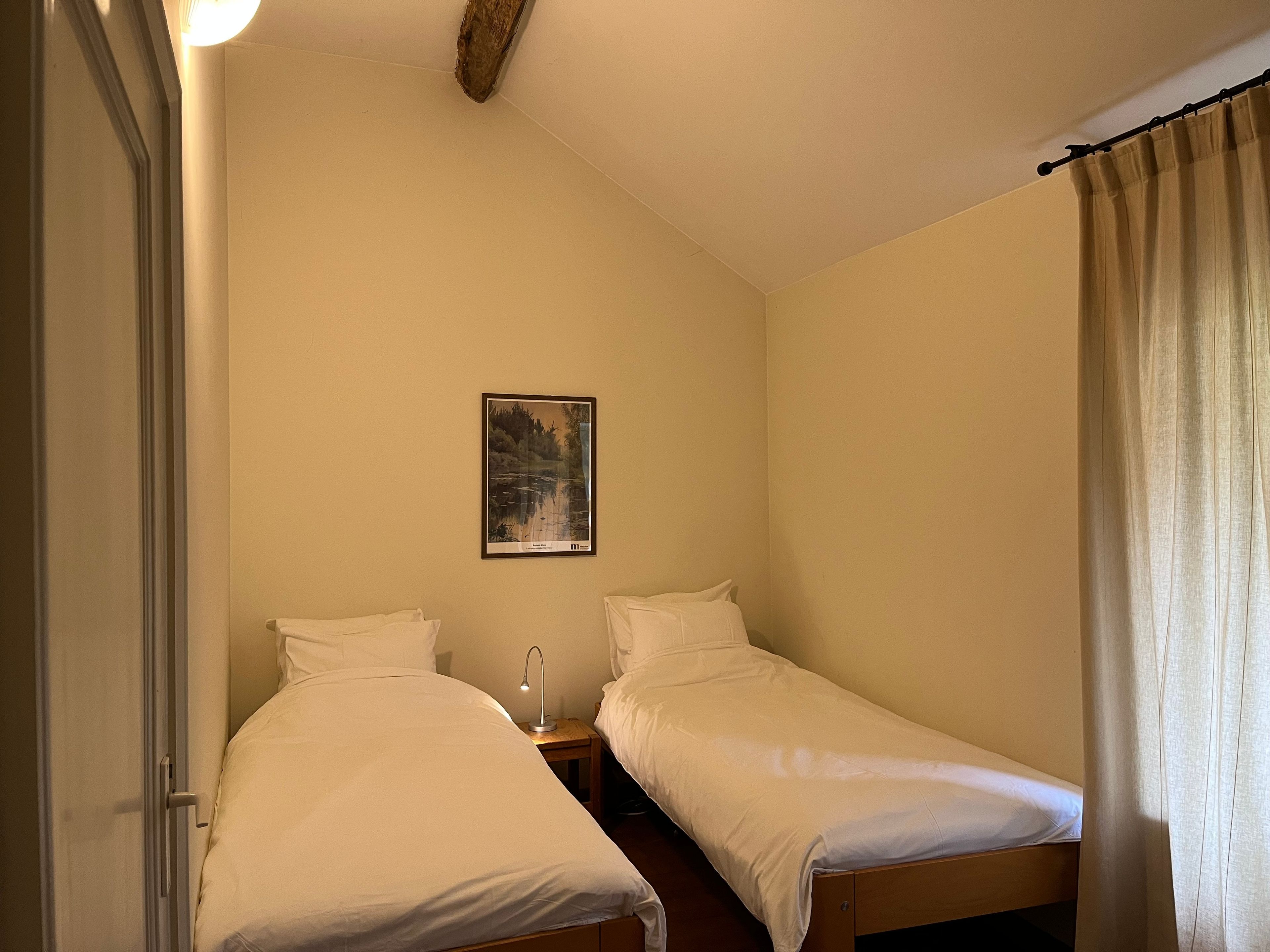 A small, single-room accommodation with two single beds, a nightstand with a lamp, a framed picture on the wall, and a door.