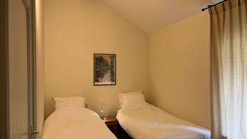 A small, single-room accommodation with two single beds, a nightstand with a lamp, a framed picture on the wall, and a door.