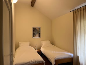 A small, single-room accommodation with two single beds, a nightstand with a lamp, a framed picture on the wall, and a door.