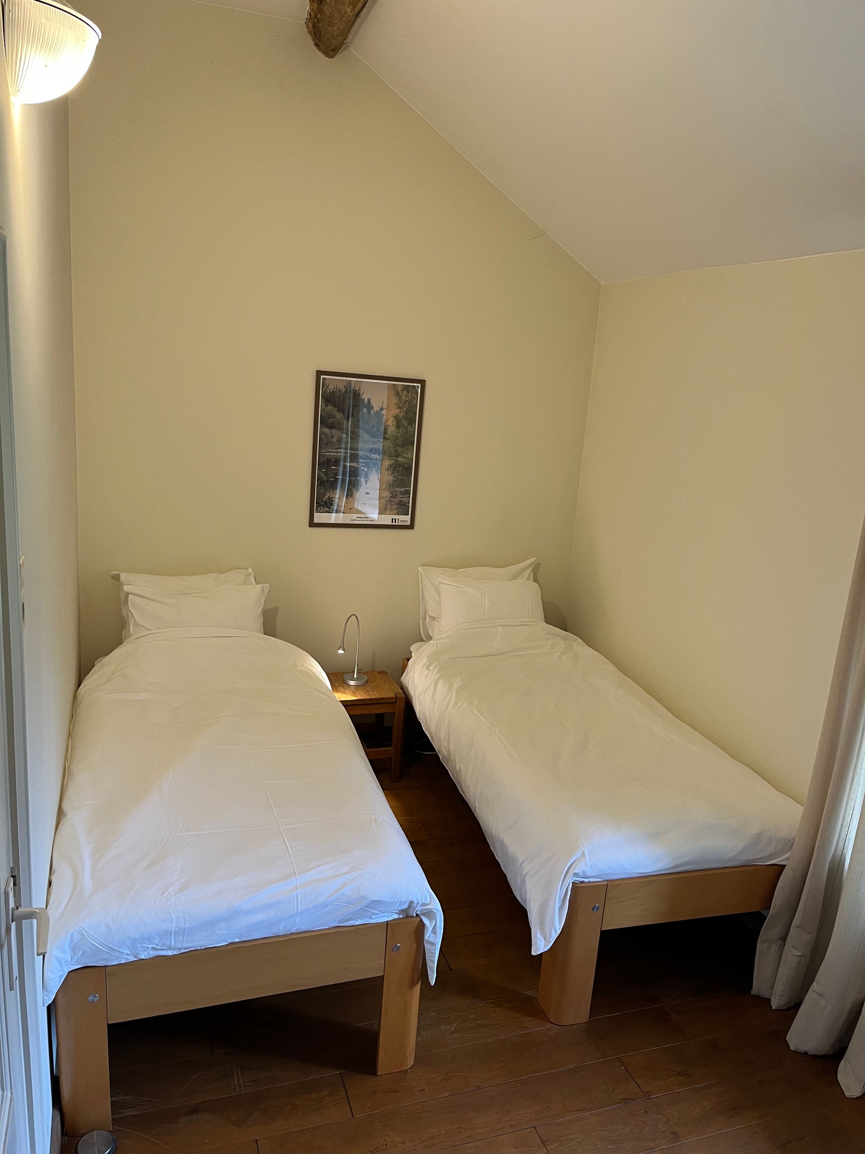 A room with two single beds, a nightstand with a lamp, a framed picture on the wall, and a wooden floor.