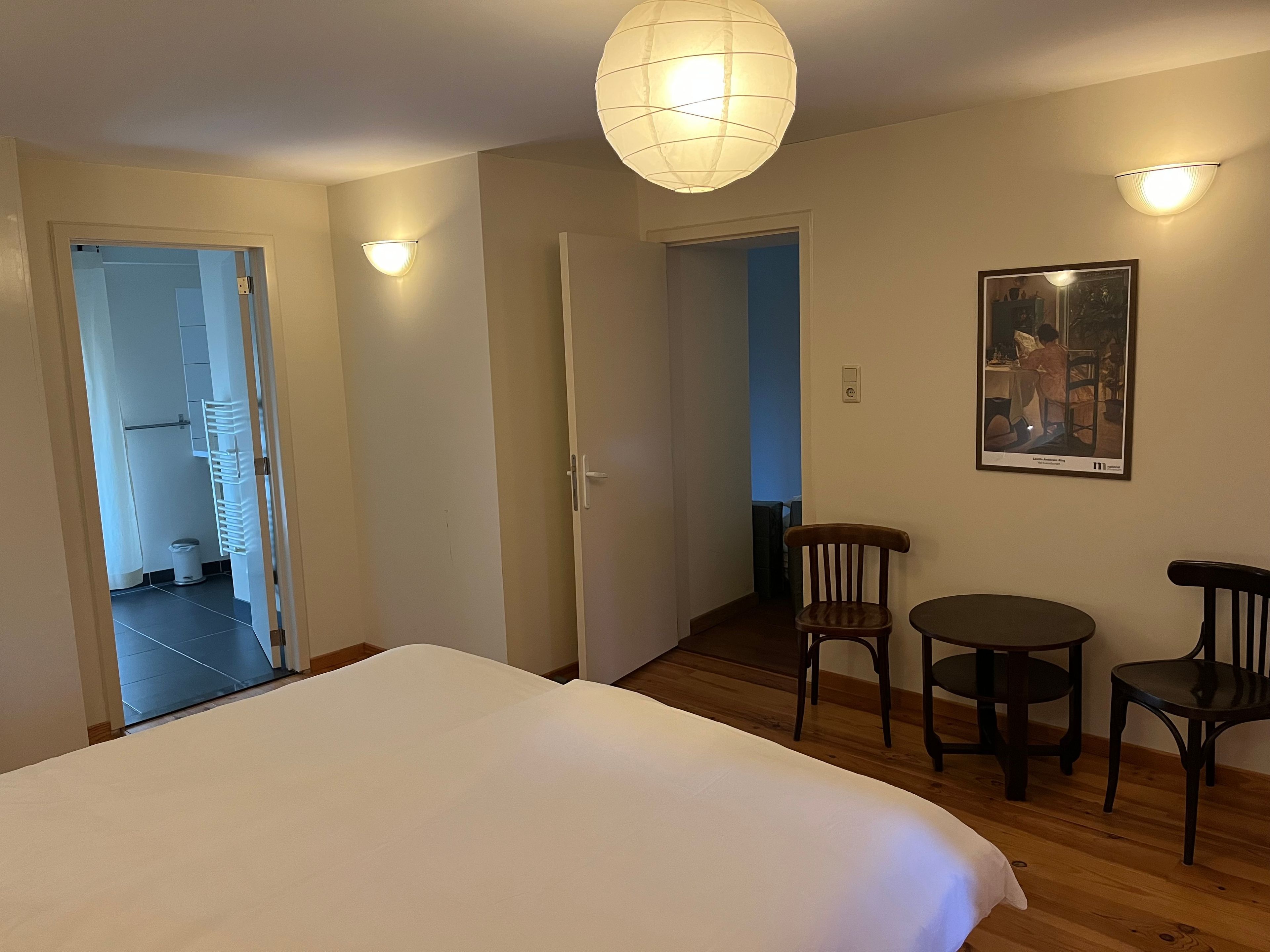 Habitación de hotel con una cama, una mesita, dos sillas, una lámpara de pared y una puerta que da a otra habitación.